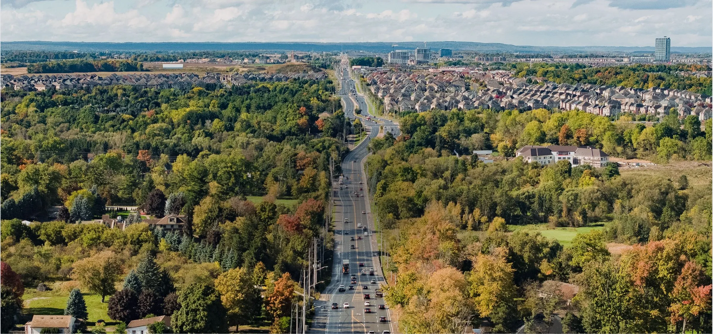 Photo of a wide road through autumn woods and suburbs.